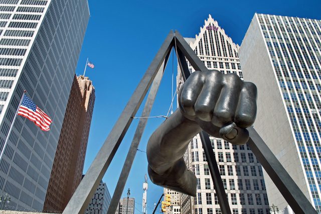 Giant metallic fist hangs within a triangular frame, surrounded by tall buildings and American flags in a cityscape under a clear blue sky.