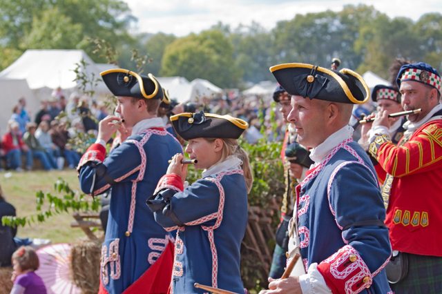 Musicians play fifes in historical uniforms at an outdoor event, surrounded by blurred spectators and tents on a sunny day.