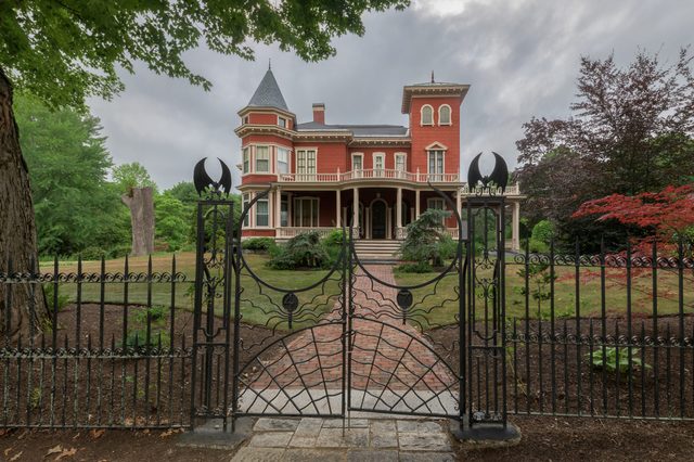 Ornate wrought iron gate stands closed, leading to a red Victorian-style house surrounded by lush green trees and a brick walkway.