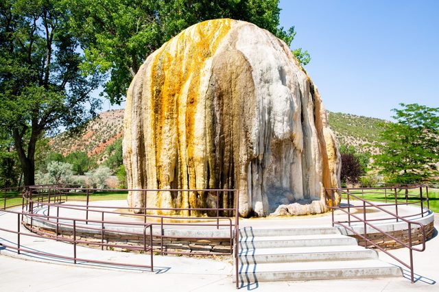 Rocky limestone formation sits surrounded by railings, flanked by trees and hills, under a clear blue sky in a landscaped park setting.
