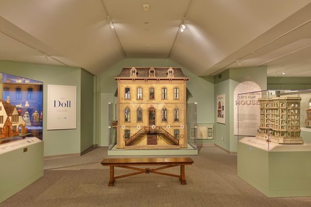 Dollhouse displayed behind glass case, inside museum room. Text reads "The Doll Gallery" and "Let's Play House" on nearby walls. A wooden bench is placed in front.