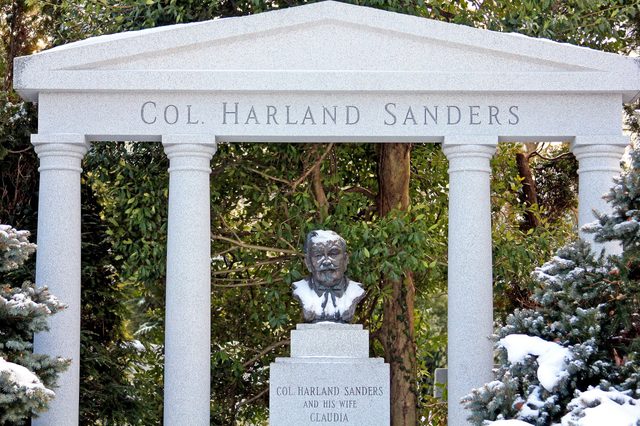 A bust rests on a pedestal amid columns and greenery, beneath the inscription "COL. HARLAND SANDERS." Snow lightly covers the monument and surroundings.