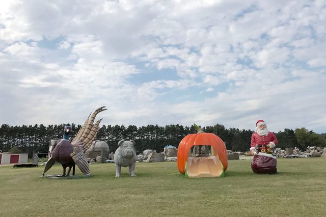 Statues stand on grass: a pheasant, bulldog, pumpkin slide, and Santa, with trees and cloudy sky in the background.
