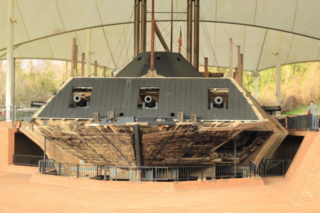 Shipwreck displayed in a covered outdoor area, featuring cannons and a wooden, weathered structure. Visitors observe the historical exhibit surrounded by railings and foliage.