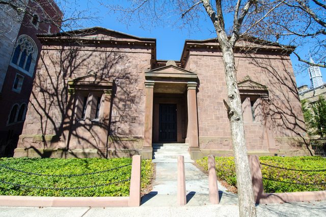 A brown stone building stands, casting shadows from surrounding trees, with a prominent black door, steps, and minimal greenery under a clear blue sky.