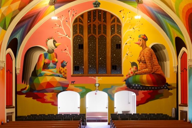 Vivid murals depict abstract figures sitting, surrounded by colorful patterns in a church with high ceilings and wooden benches below.