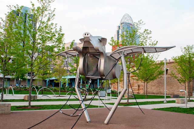 Metal pig sculpture with wings stands as playground equipment in a park, surrounded by trees and city buildings in the background.