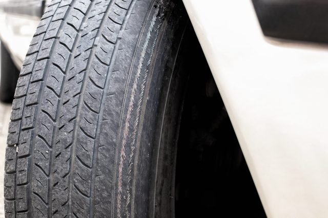 A damaged car tire shows visible wear, parked on a concrete surface next to a white vehicle body.