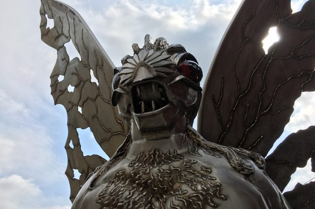 Metallic creature statue stands still, displaying intricate wings and fierce expression against a cloudy sky backdrop.