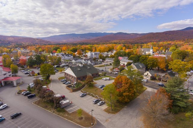 Buildings sit surrounded by colorful autumn trees, with mountains in the background and parked cars in the foreground under a partly cloudy sky.