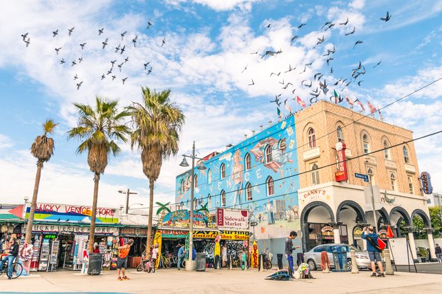 Buildings feature murals and shops bustling with people; birds fly across a blue sky. Palm trees and street signs enhance the lively street scene.