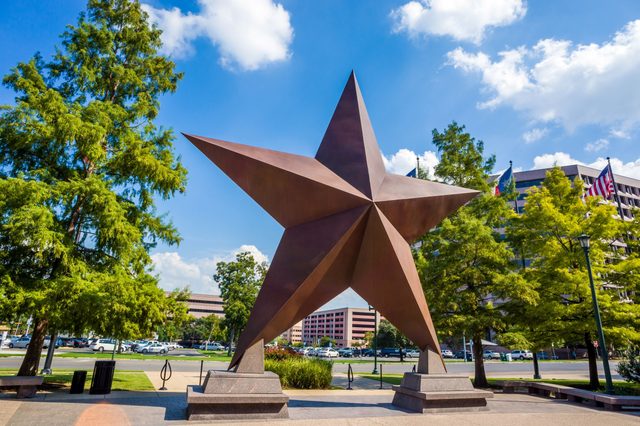 Bronze star sculpture stands prominently in an urban park with trees, flagpoles, and buildings under a clear blue sky with scattered clouds.