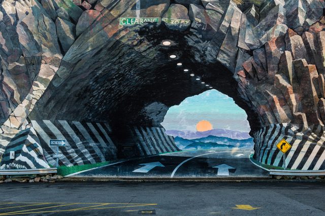 Tunnel mural depicts a road extending towards a sunset, surrounded by rocky walls. Street signs indicate "CLEARANCE" and "ONE WAY" in a parking lot.