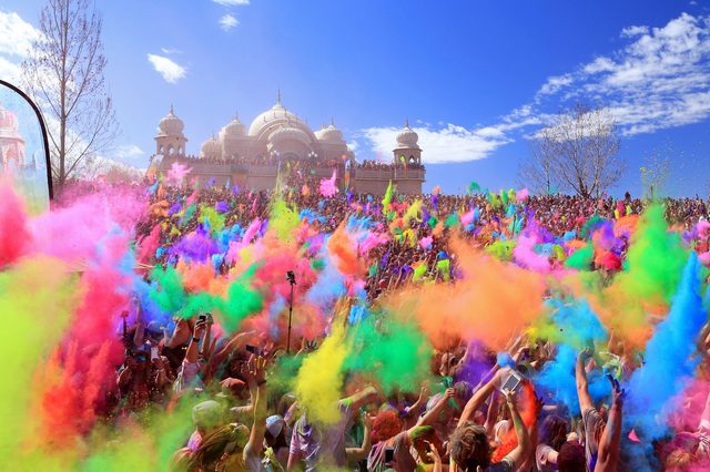 Crowd throws colorful powders, creating vibrant clouds in front of a grand building with domes on a sunny day.