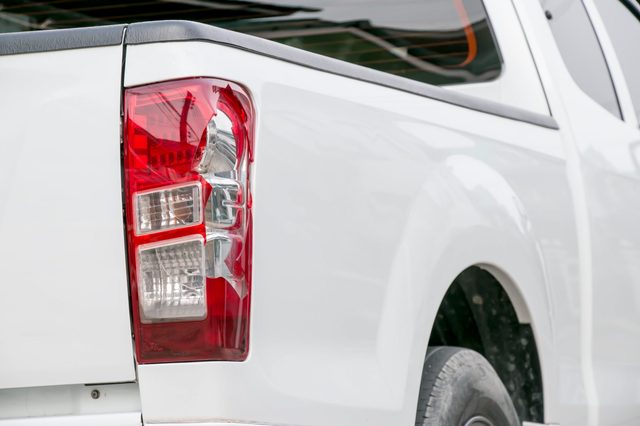 A white pickup truck parked; its rear taillight is visible, surrounded by a reflective glossy surface under indirect daylight.