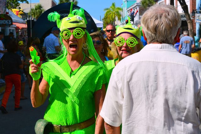 Two people in bright green alien costumes walk excitedly in a crowded street, holding water guns on a sunny day.