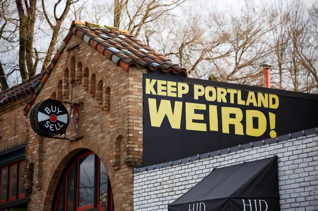 Sign displays "KEEP PORTLAND WEIRD!" on a brick building with a 'BUY SELL' vinyl record sign nearby, surrounded by leafless trees.