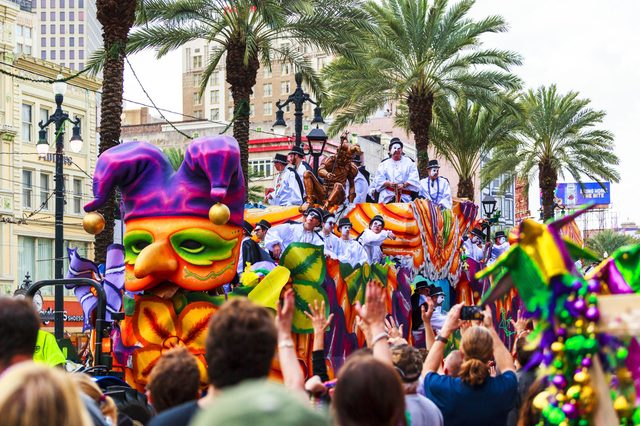 Colorful parade float passes through a lively street, featuring people in costumes surrounded by palm trees and buildings.