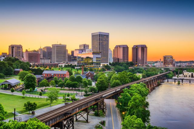 City skyline at sunset features tall buildings behind lush green parks, with a river and bridge in the foreground.