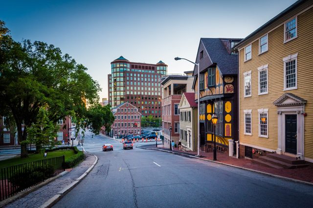 Street descends between historic and modern buildings; cars drive downhill. Trees line the sidewalk under a clear sky at dusk, highlighting architectural contrast.