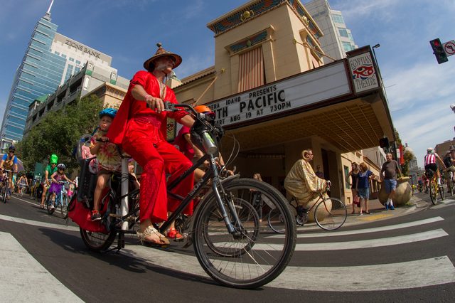 A person in a vibrant red costume rides a bicycle with a child in a city parade, passing a theater advertising "South Pacific" on August 19.