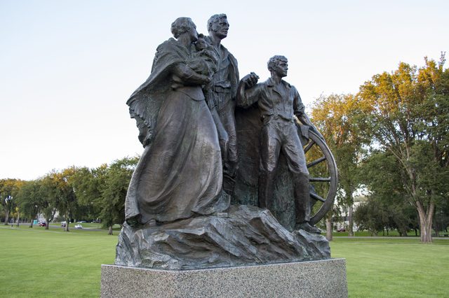 Bronze statue depicts a pioneering family standing resolutely, surrounded by grassy parkland with trees in the background.