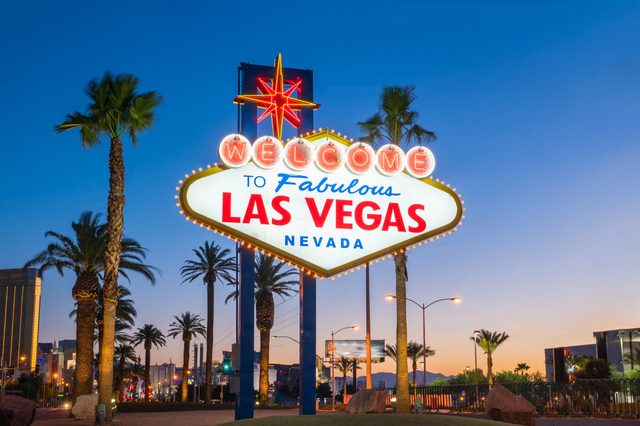 Sign displays "Welcome to Fabulous Las Vegas, Nevada," glowing. Palm trees and city lights surround, with a twilight sky in the background.