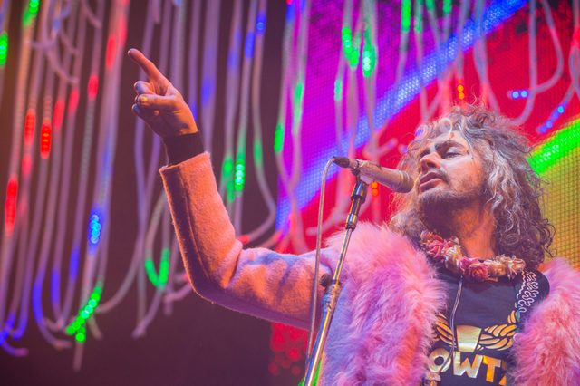 Performer gestures while singing into a microphone, wearing a colorful outfit. Bright multicolored lights and hanging decorations create a vibrant stage backdrop.