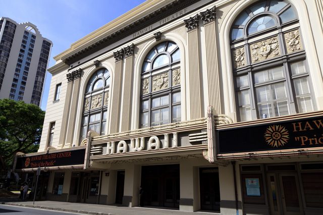 Theater building displays "HAWAII" on marquee; tall windows, ornate details. Context includes urban street, tree, and neighboring skyscraper under a blue sky.