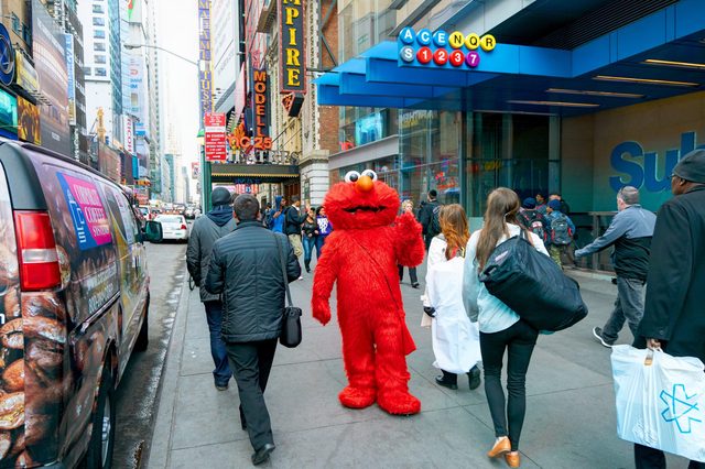 Character in red costume walks on busy city sidewalk, surrounded by pedestrians and colorful storefront signs.