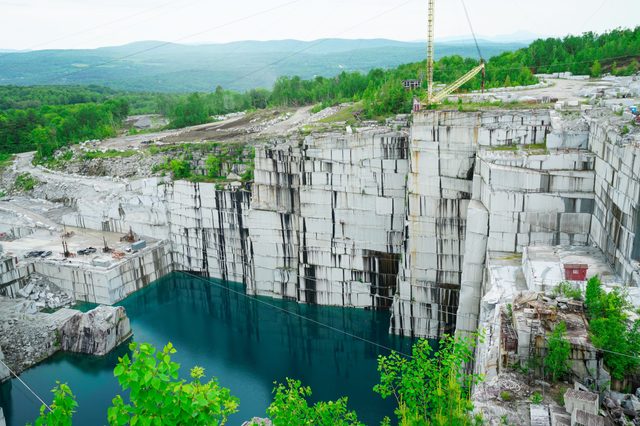 A deep marble quarry with still blue water, surrounded by lush green forest and distant hills under a cloudy sky.