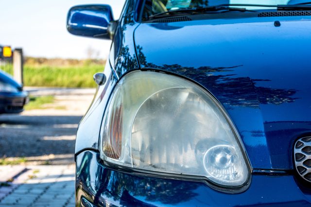 Car's left headlight reflecting trees, parked in a sunlit area with a blurred grassy background and another car visible nearby.