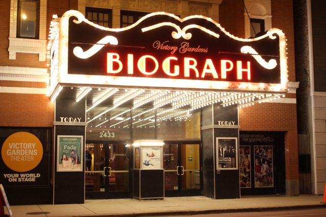 Theater entrance shines bright with marquee lights, displaying "Victory Gardens Biograph" in a city setting, posters line walls, brick building backdrop.