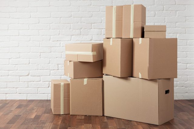 Cardboard boxes stacked, secured with tape, against a white brick wall on a wooden floor.