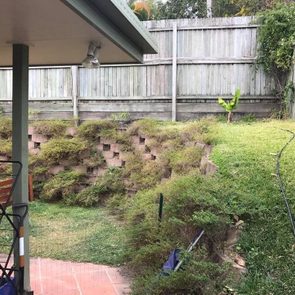 Backyard garden displays terraced plants on a slope beside a tiled patio with a wooden fence surrounding the area.