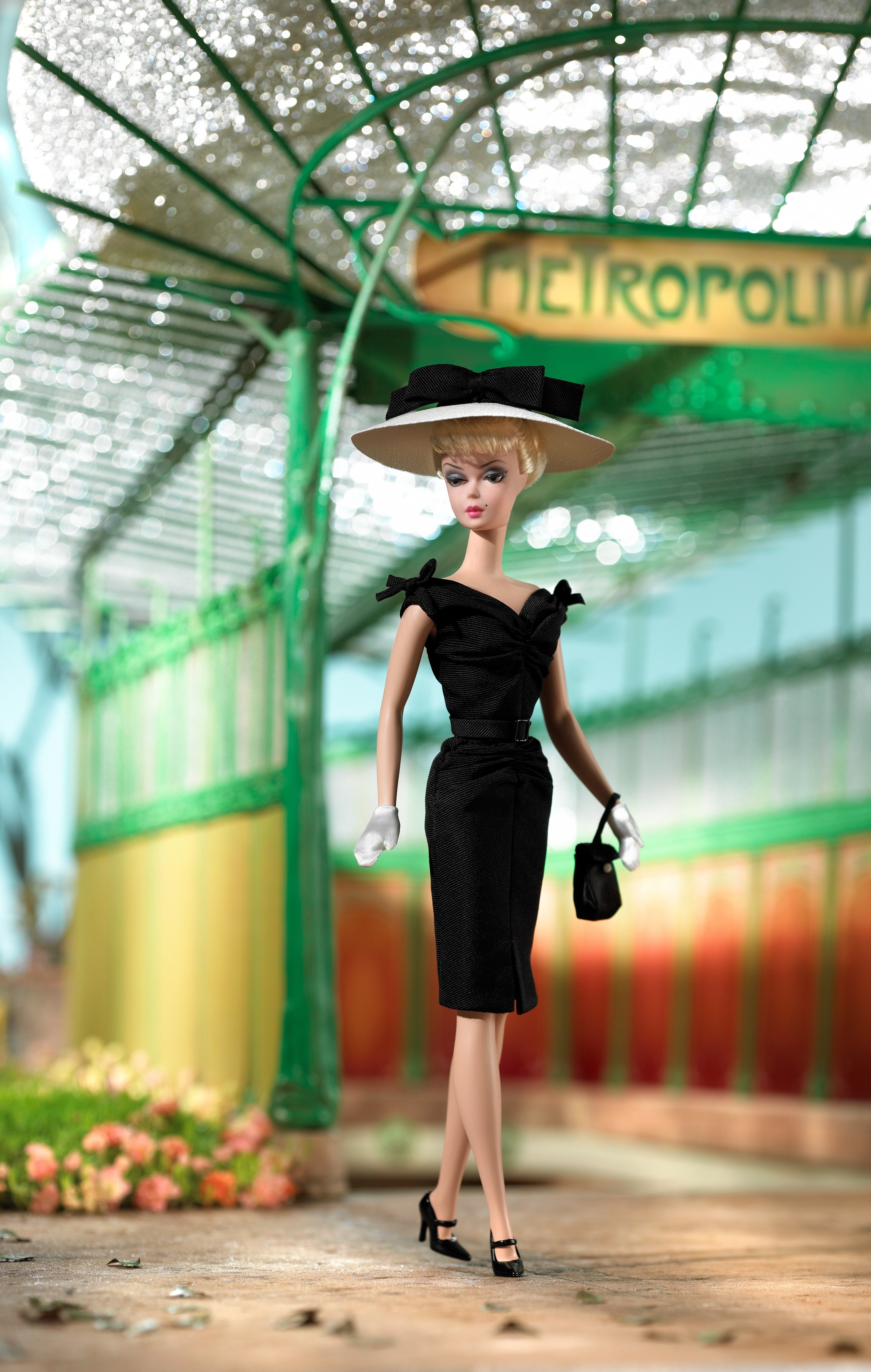 A doll in a black dress and hat stands elegantly, holding a purse in a stylized, vintage metro station setting.