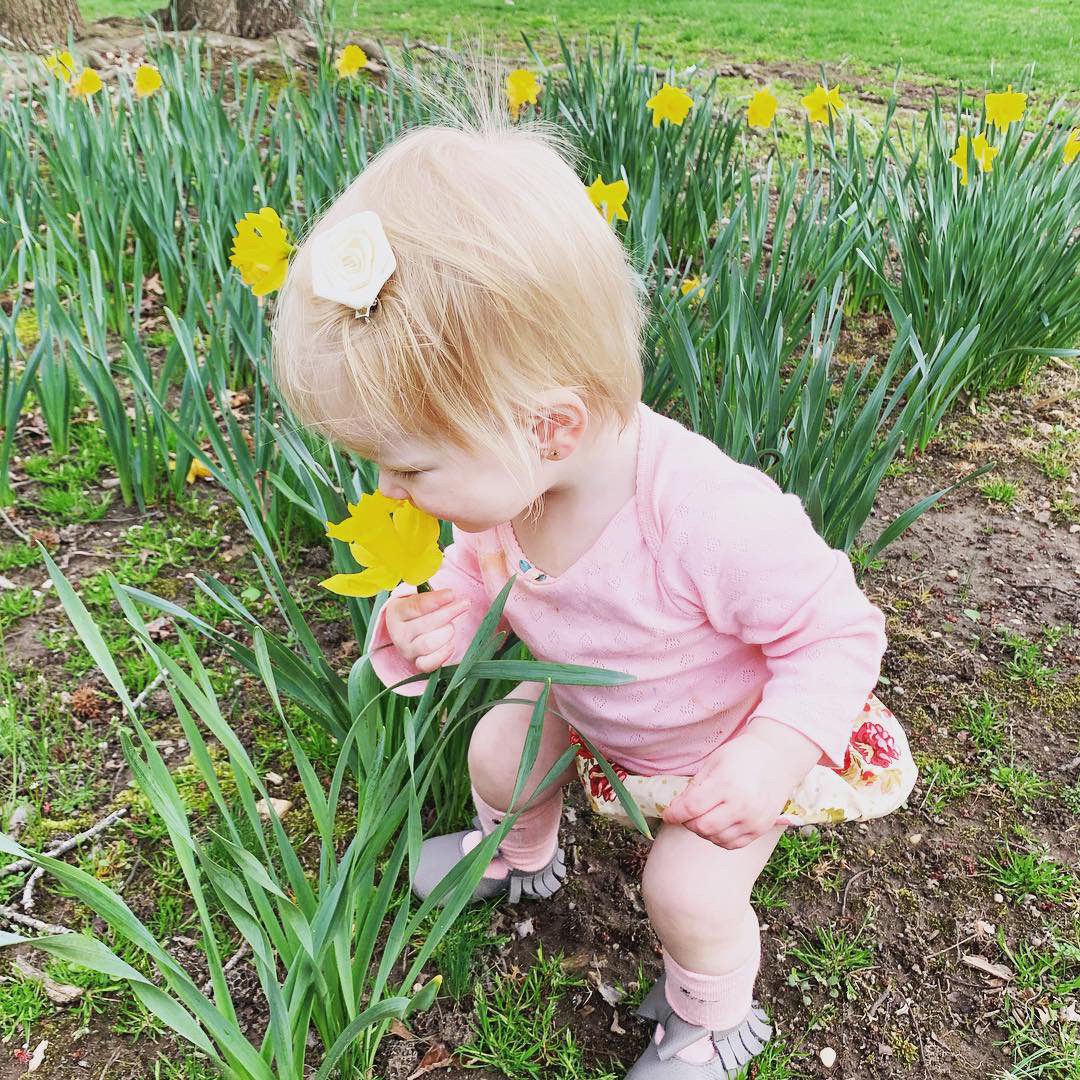 Child sniffs yellow daffodil, crouching in a grassy field surrounded by more daffodils.