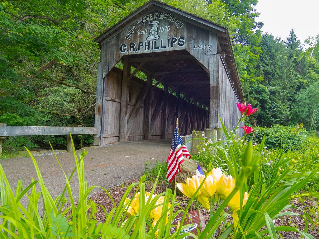 Covered wooden bridge stands amidst vibrant flowers and flags, surrounded by lush green trees. Text reads: â€œBUY YOUR DRUGS OF C.R. PHILLIPS Built 1876 SALEM O.â€
