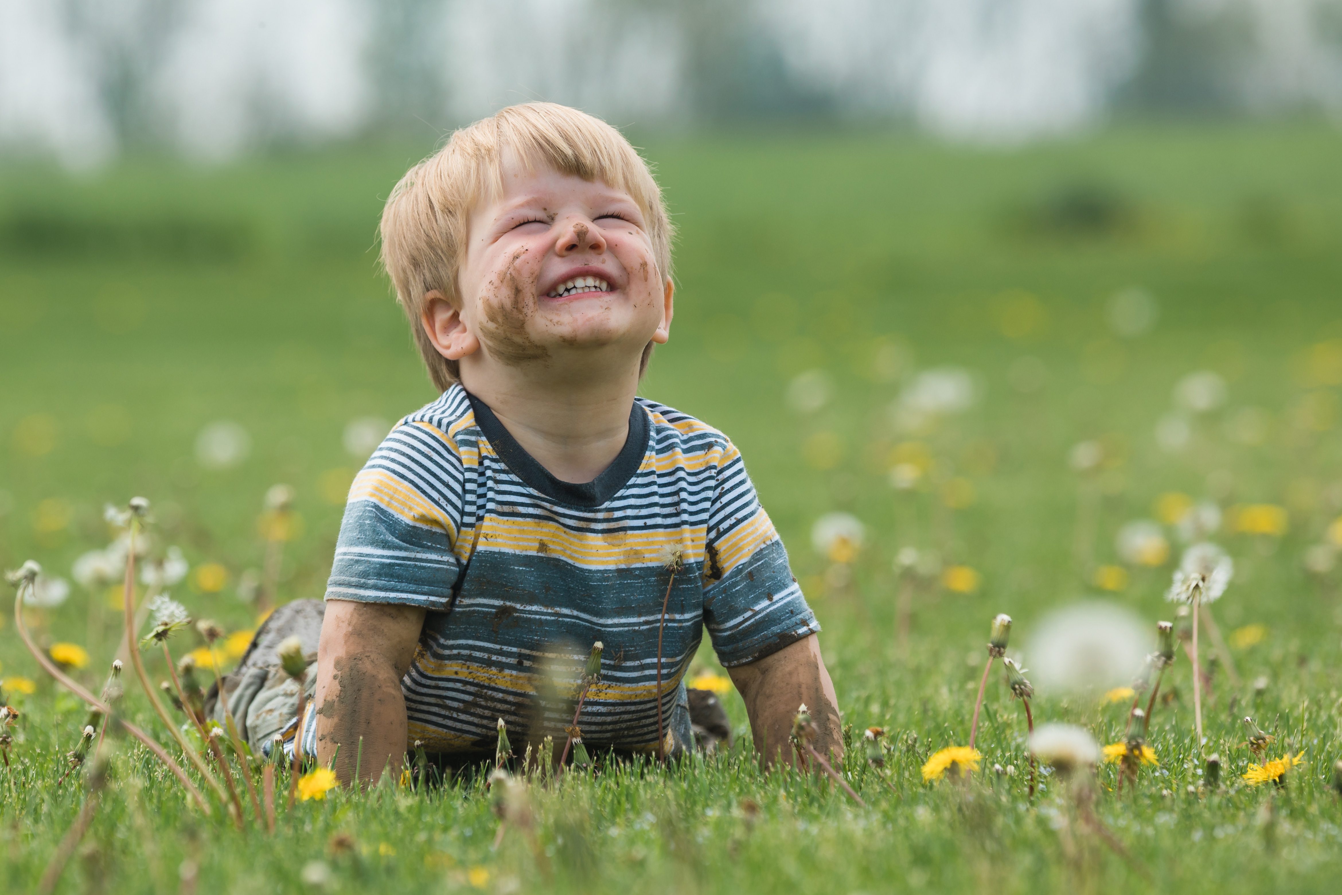 Child smiles while sitting on grassy field, surrounded by dandelions, with a joyful expression and dirt on face and arms.