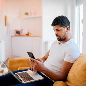 Man sits on a yellow couch, using a smartphone and laptop, in a bright living room with a cup of coffee on a side table.