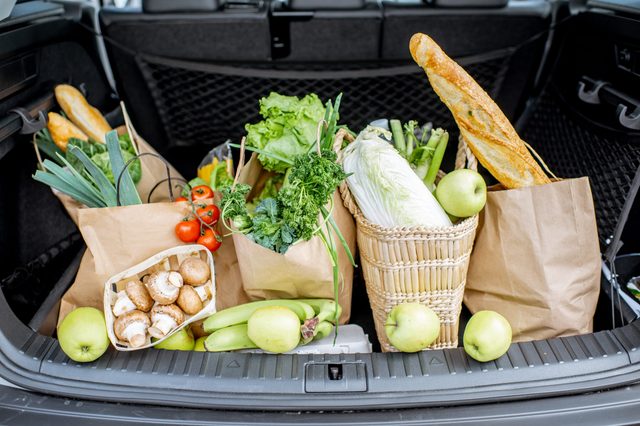 Groceries sit in the car trunk, with vegetables and baguettes in paper bags, surrounded by apples, mushrooms, and bananas.