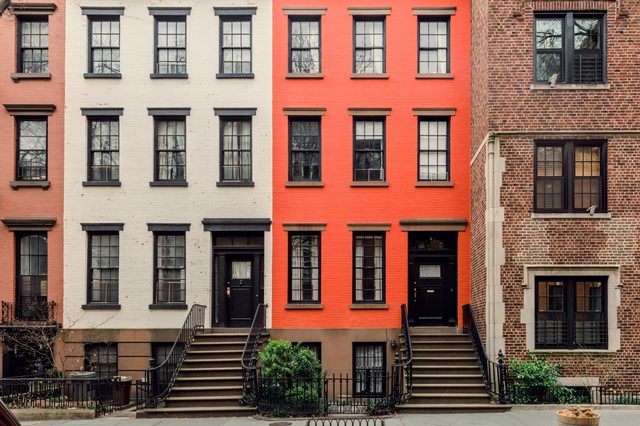 Three townhouses stand aligned; middle one painted red, flanked by white and brown facades. Steps lead to black doors, greenery surrounds entrances.