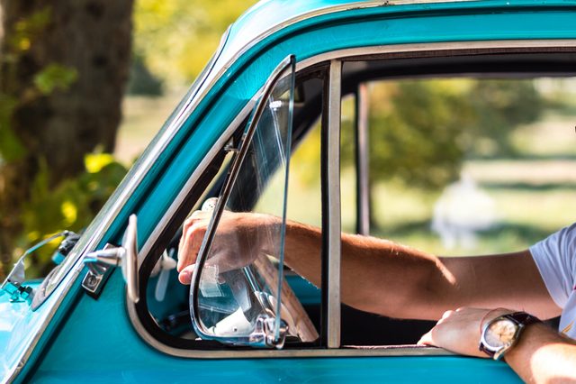 A person's arm rests out of an open window of a vintage teal car in a sunny, green outdoor setting.
