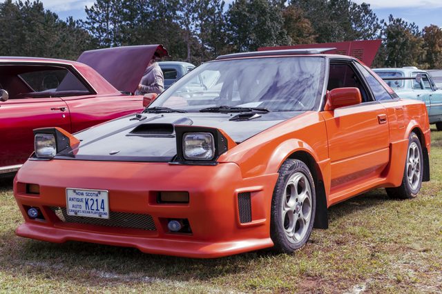Orange sports car parked outdoors, showcasing pop-up headlights and Nova Scotia license plate "K214" amid other vintage vehicles in a wooded setting.