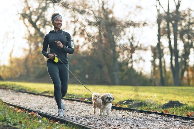 Woman jogging with a small dog on a leash, surrounded by a grassy park with trees in the background.