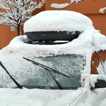 Car covered in snow; windshield wipers raised; snow on ground and tree branches; parked in front of orange building; visible unit number 20104.