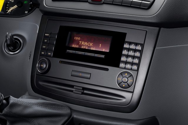 Car stereo displays "TRACK 1," surrounded by control buttons, within a dark, textured dashboard. Gear shift and ignition visible nearby.