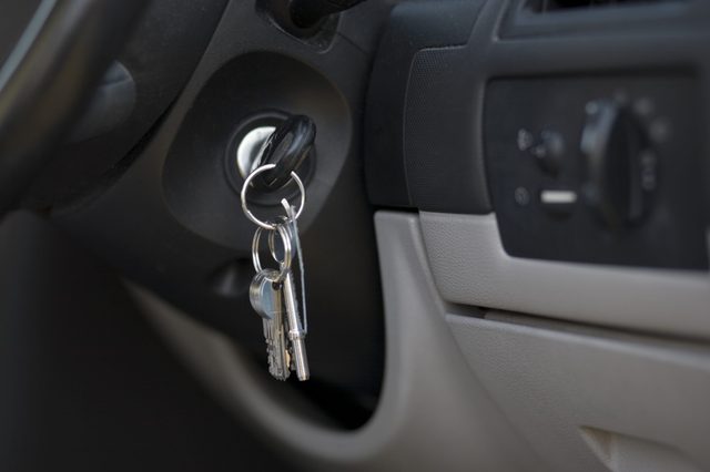 Keys hang in a car ignition, surrounded by a dashboard and steering wheel in a vehicle interior.