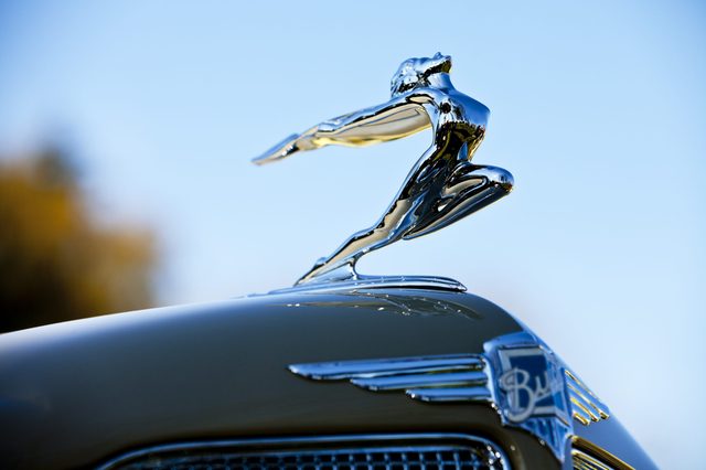 Chrome hood ornament depicts a leaping figure, mounted on a car. Background shows a clear blue sky with a hint of trees.