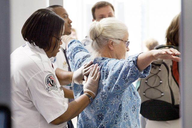 Security officer performs a pat-down on a woman in a floral blouse at an airport security checkpoint. Others wait in line nearby.